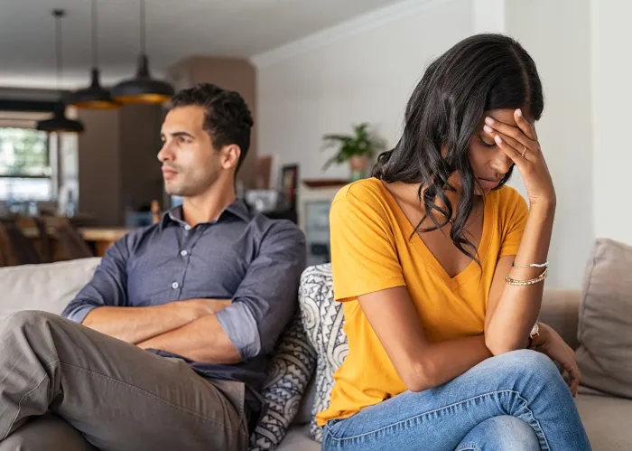 Middle eastern young couple sitting on couch after a fight. Sad indian woman sitting with hand on head after quarrel with boyfriend at home. Angry latin couple ignoring each other on the sofa, having relationship troubles.