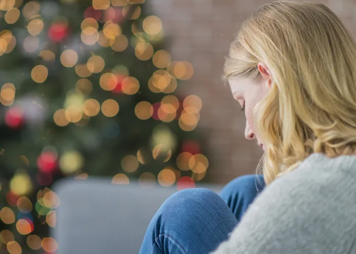 A Caucasian woman is indoors in her living room. There is a Christmas tree in the background. The woman is wearing warm clothing. She is sitting on the couch and looking sad because she is alone on Christmas day.