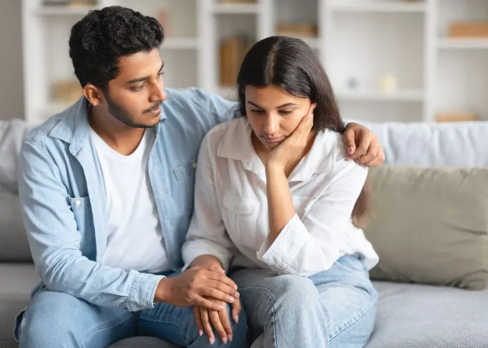 Caring young indian husband next to wife, lovingly attempting to soothe and console woman, providing comforting presence, sitting on sofa at home